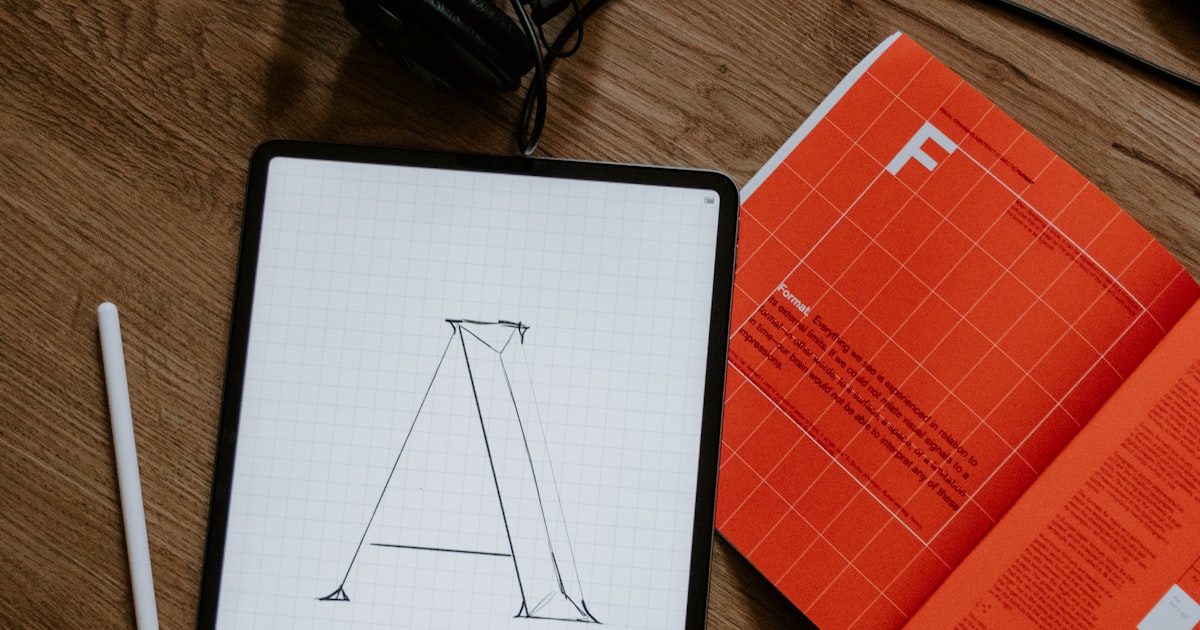 Tablet con boceto de letra tipográfica geométrica sobre papel cuadriculado junto a revista de diseño tipográfico roja sobre escritorio de madera
