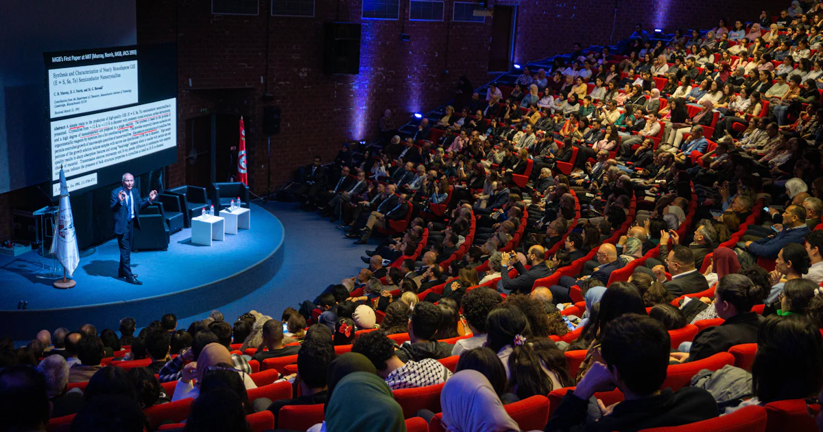 Speaker presenting to a large audience in an auditorium, seen from the back of the room