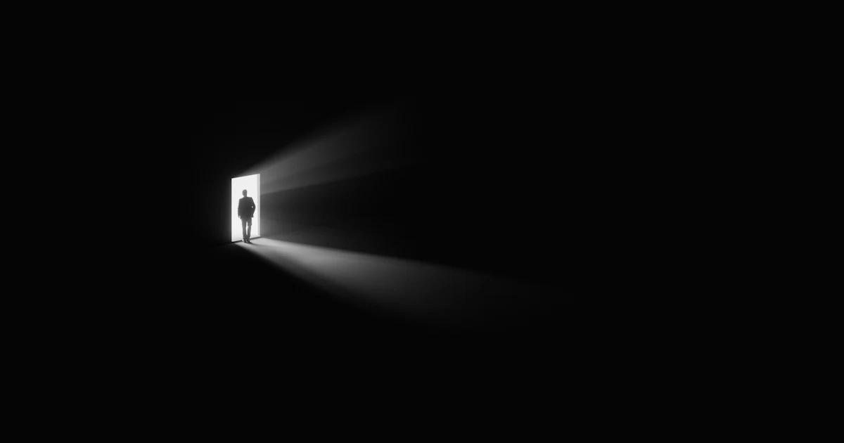 Silhouette of a person standing in a lit doorway, between the darkness of a room and the light outside