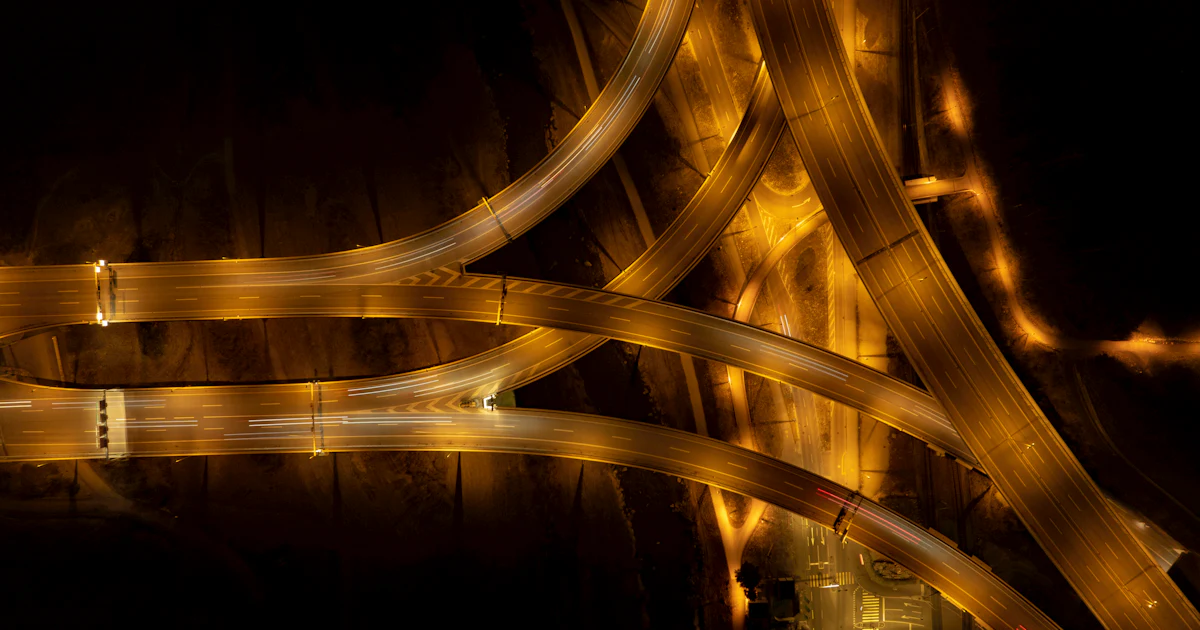 Vista aérea nocturna de un cruce de autopistas con las luces de los vehículos formando trazos de distintos caminos